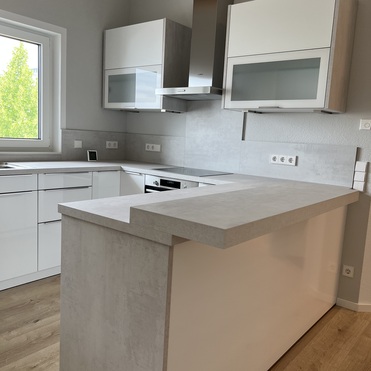 Eine helle Küche nach Maß mit weißen und hellbraunen Schränken, grauem Fliesenspiegel und einer Kochinsel, beleuchtet durch Tageslicht vom Fenster.