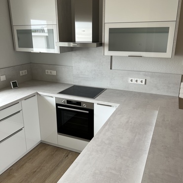 Maßgefertigte Küche mit weißer Einrichtung, eingebautem Ofen und Kochfeld auf einem hellen Holzboden, unterstrichen durch ein großes Fenster.