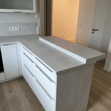 Ansicht einer Kücheninsel nach Maß mit weißer Front und steinfarbener Arbeitsplatte in einem modernen Kücheninterieur mit Holzboden.