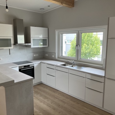 Maßgefertigte Küche mit heller Farbgebung, modernen Schränken und Geräten, einer Kochinsel und natürlichem Licht durch das Fenster.