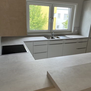 Ansicht einer maßgefertigten Küche mit weißer Schrankfront, eingebautem Cerankochfeld und einem Fenster, das auf grüne Bäume blickt.