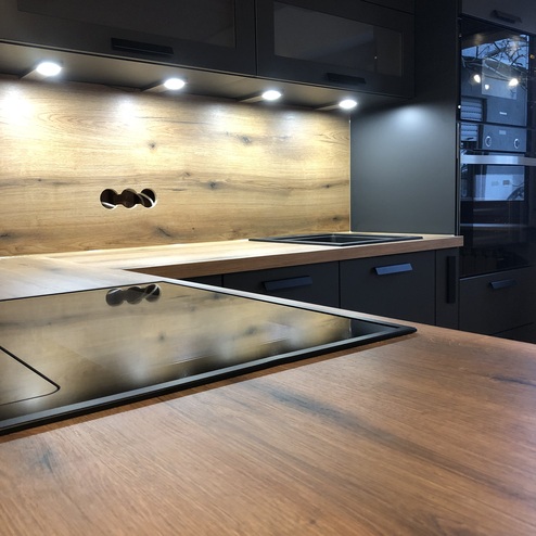 Eine Küche nach Maß mit dunklen Schränken, integrierten LED-Leuchten und einer Arbeitsplatte in Holzoptik, die Wärme und Modernität ausstrahlt.