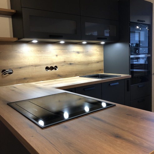 Detailansicht einer Küche nach Maß mit eleganten dunklen Schränken, Holzakzenten und modernen Kochfeldern, beleuchtet durch Unterbauleuchten.