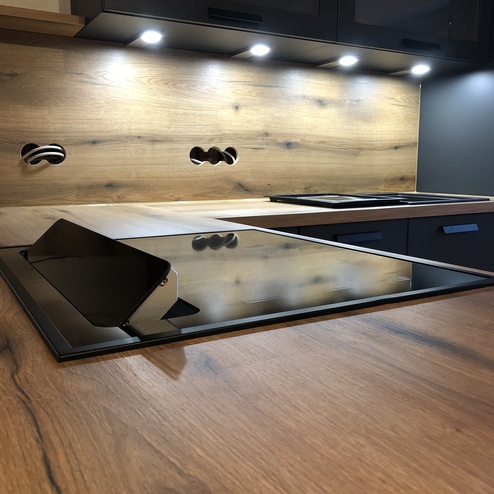 Perspektivische Ansicht einer Küchenarbeitsfläche nach Maß mit Holzoptik, modernen schwarzen Einbaugeräten und effektvoller Unterbaubeleuchtung.