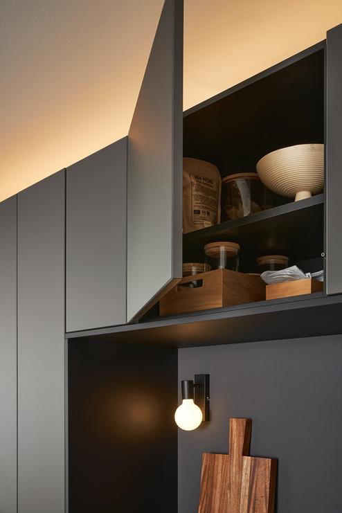 Ge&ouml;ffneter K&uuml;chenoberschrank mit Holzboxen, Vorratsgl&auml;sern und dekorativer Schale &uuml;ber moderner Wandbeleuchtung