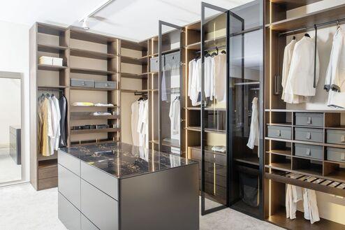 Gro&szlig;z&uuml;gige Ankleide nach Ma&szlig; mit Mittelinsel, Glasfronten und Regalsystemen in dunklem Holzton.